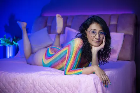 Smiling woman in a colorful rainbow bodysuit relaxing on a bed with moody blue and purple lighting.