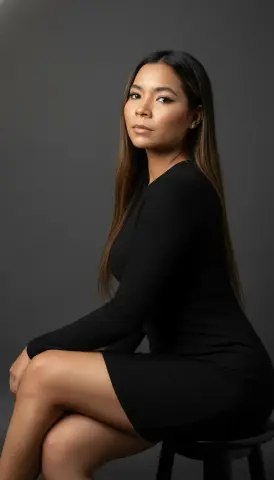 XinemaBonet poses sitting on a stool, wearing a tight black long-sleeved dress with her long brown hair down against a grey background.
