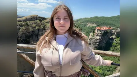 Young woman in a beige jacket smiling on a wooden balcony overlooking the Meteora monasteries in Greece.