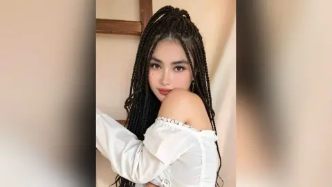 Young woman with long dark braids wearing a white off-the-shoulder blouse in a soft, bright indoor setting.
