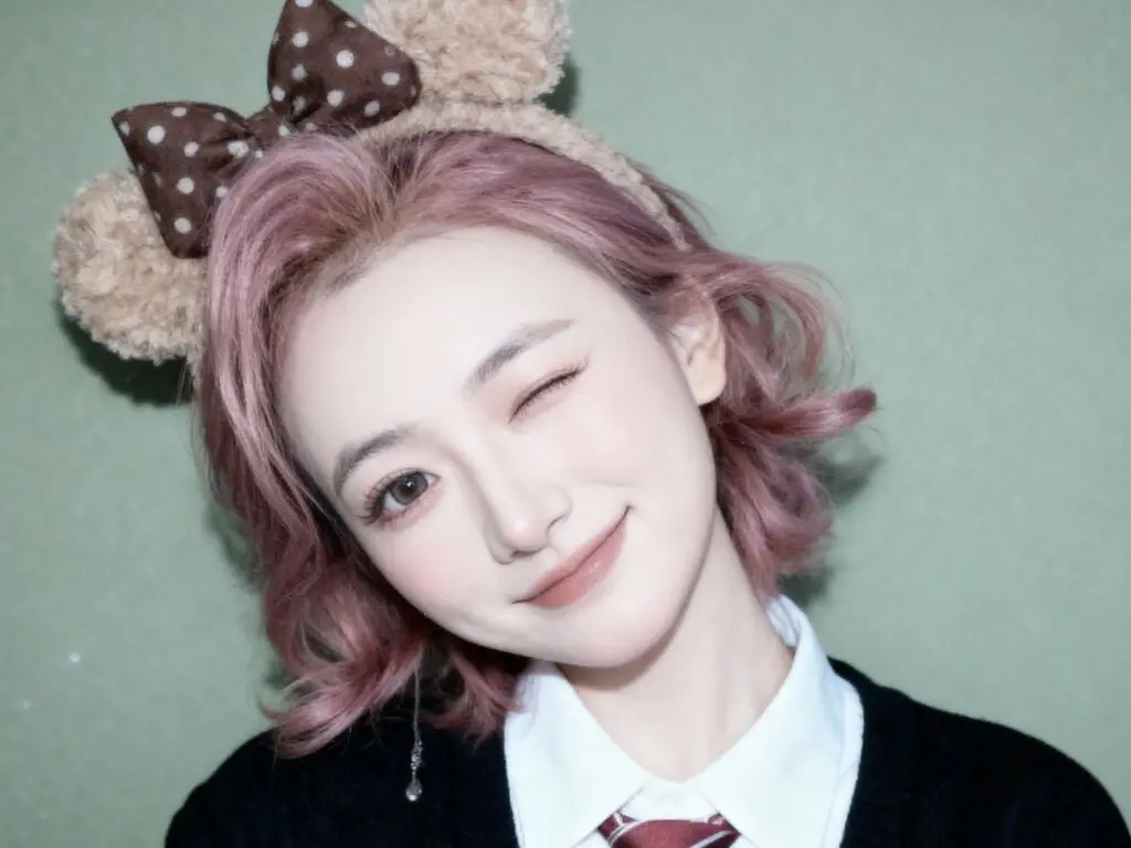Smiling young woman with pink hair wearing a brown polka dot bear ear headband against a green background.