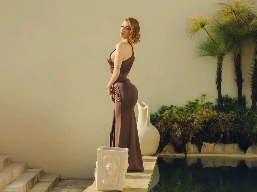 Elegant woman in a brown evening gown standing by a luxury pool in a serene outdoor garden setting.