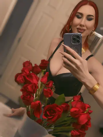 Redheaded woman in a black crop top holding red roses for a romantic mirror selfie.