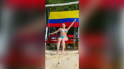 Cheerful woman in a blue bikini sitting on a wooden swing in front of a large Colombian flag on a sunny beach.