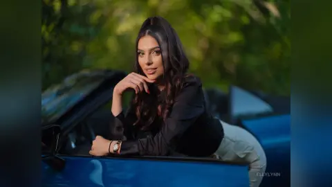 Elegant woman in a black blouse leaning on a blue convertible car in a lush green outdoor setting.
