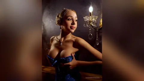 SashaNavikova poses in a blue lace corset, pulling down the top to expose her breasts near a lit wall lamp.