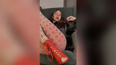 Woman in black top and red patterned tights relaxing on a green sofa with a moody, stylish aesthetic.