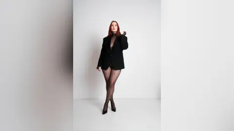 AryaSin poses in a black blazer and sheer black tights against a plain white background, looking directly at the camera with her red hair down.