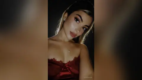 ArianaRoux poses in a red lace bustier, looking at the camera with long dark hair and heavy eye makeup against a dark background.
