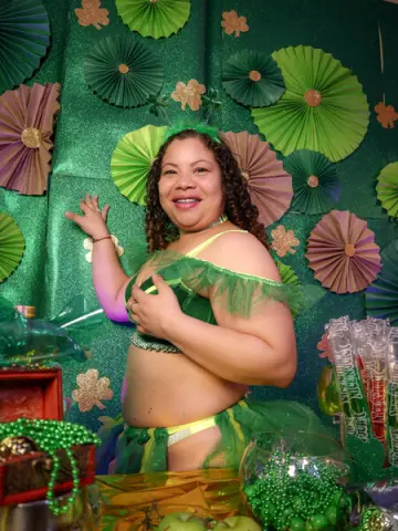 Smiling woman in a green festive outfit posing against a green paper fan backdrop at a party.