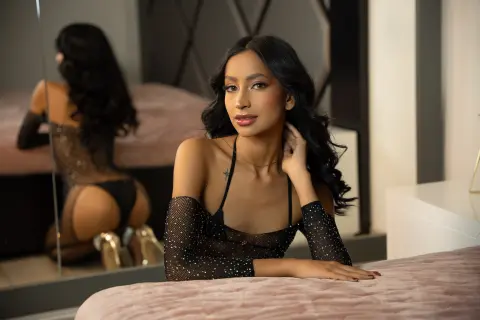 Elegant woman in a black sparkly sheer top relaxing on a bed in a modern bedroom setting.
