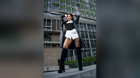 Stylish woman in a black trench coat and white shorts posing confidently on a modern urban rooftop.