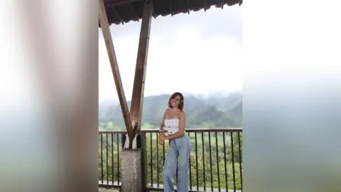 AmelieLautrec stands on a balcony overlooking a lush, green mountain landscape, wearing a white tube top and light blue jeans.