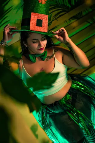 Woman in white tank top and green glittery accessories posing in a lush, moody tropical garden setting.