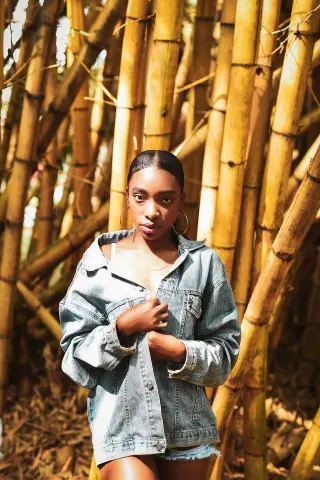 Young Black woman in a denim jacket posing serenely amidst a lush golden bamboo forest.