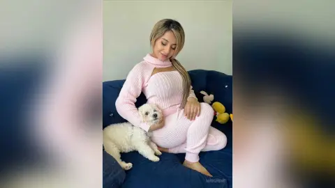 Smiling woman in a cozy pink knit sweater sitting on a blue sofa with a small white fluffy dog.