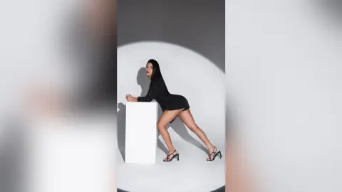 Elegant woman in a black mini dress and strappy heels posing against a minimalist grey studio backdrop.