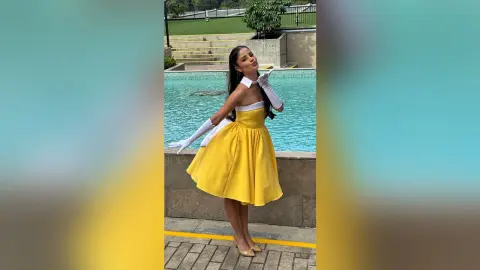 TefyRusso poses in a yellow strapless dress and white gloves by a swimming pool, looking back over her shoulder with long dark hair.