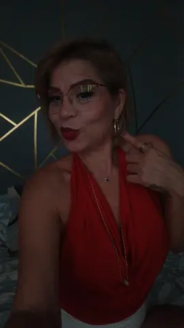 Smiling woman in a red halter top and gold jewelry posing in a moody, dark room with gold geometric decor.