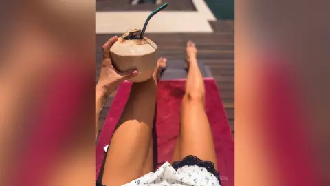 AnaMathers holds a coconut drink while relaxing with her legs extended on a red surface outdoors.