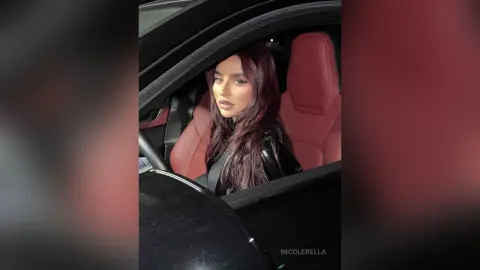 NicoleBella looks toward the camera while sitting in the driver's seat of a car with red leather seats.