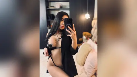 Young woman in black lingerie and an open cardigan taking a mirror selfie in a cozy bedroom setting.