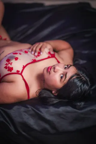 Young woman in red floral lace lingerie lying on dark satin sheets in a moody, intimate bedroom setting.