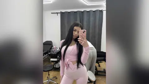 AriaVex poses in a pink long-sleeve crop top and matching pink leggings while holding a phone for a mirror selfie.