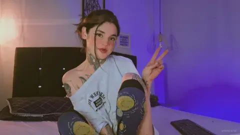 Smiling tattooed woman in a white t-shirt and patterned leggings making a peace sign in a purple lit bedroom.