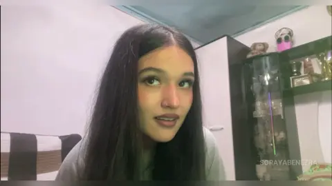 SorayaBenezra with long dark hair and a nose ring, wearing a grey top in a room with a black shelf and white walls.