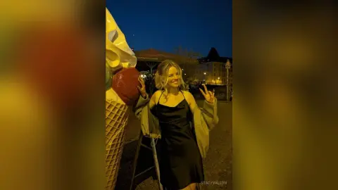 Smiling blonde woman in a black slip dress and grey cardigan posing by a giant ice cream cone at night.