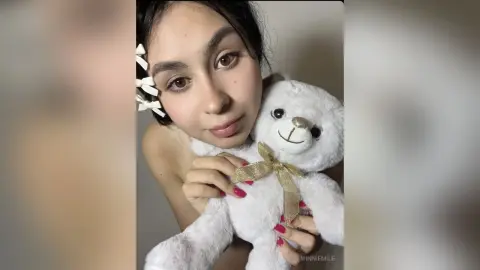 MinnieMile poses close up, holding a white teddy bear against her bare chest and shoulder, looking at the camera with dark hair styled with white bows.