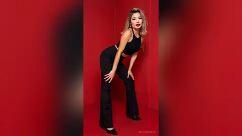 Stylish blonde woman in a black crop top and flared trousers posing against a vibrant red background.