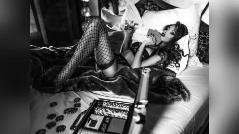 EvelynMills lies back on a bed wearing black lace lingerie and fishnet stockings, holding a small object near her chest.