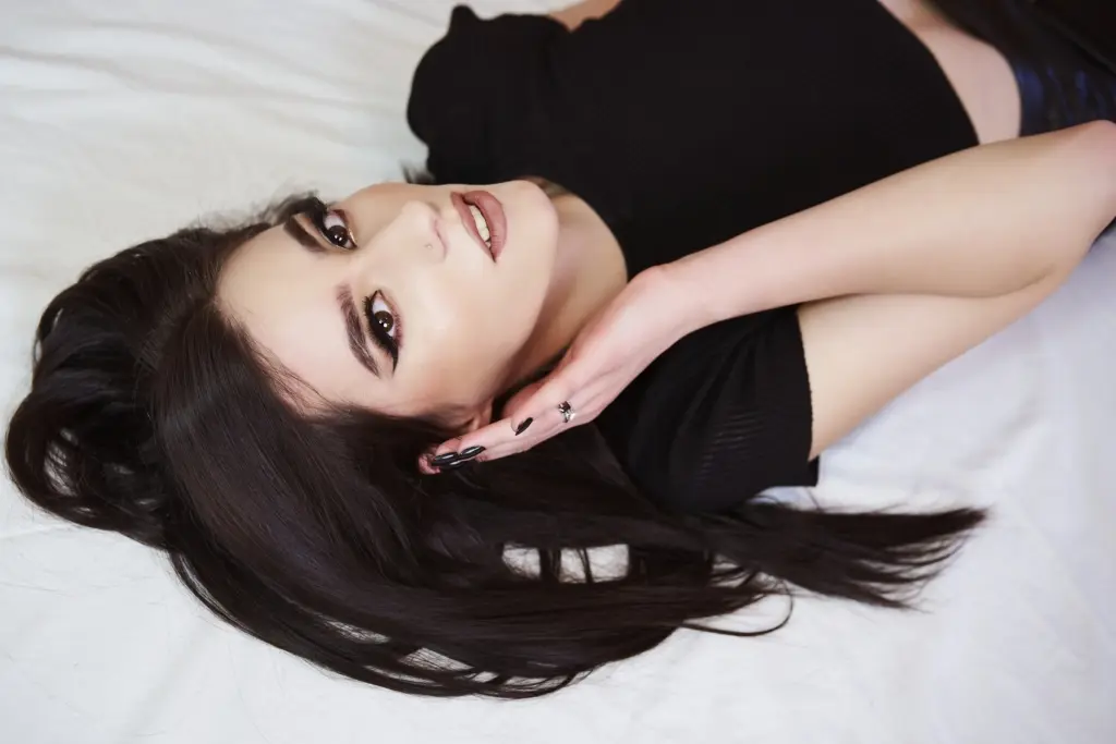 AmeliaJune lying on a white surface with her long dark hair spread out, wearing a black top and looking directly at the camera.