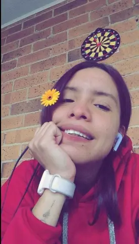 Smiling young woman in a red hoodie with a yellow flower in her hair, relaxing in a cozy room with a brick wall.
