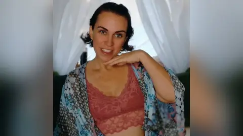 Smiling woman in a pink lace camisole and floral blue robe relaxing in a bright, airy bedroom setting.