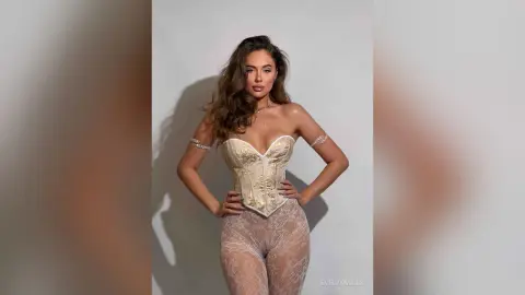 EvelynMills poses hands on hips wearing a white corset and sheer lace tights against a plain grey background.