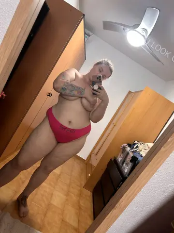 Curvy woman in pink underwear posing for a mirror selfie in a modern bedroom setting.