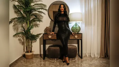 CassyeCross stands behind a wooden console table wearing a tight black long-sleeved dress, posing in front of a round mirror and a lamp.