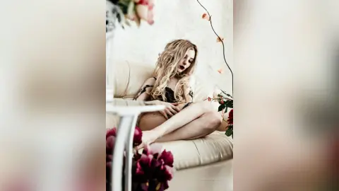 Elegant blonde woman in a floral dress relaxing on a white sofa in a bright, dreamy indoor setting.