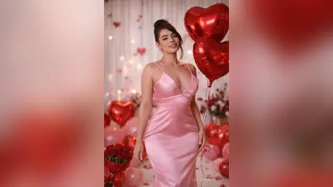 Elegant woman in a pink satin slip dress holding red heart balloons in a romantic celebratory setting.