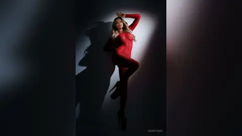 Anastasia poses in a red long-sleeved bodysuit with her arms raised against a dark background.