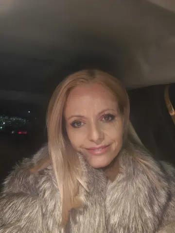 Smiling blonde woman in a cozy grey faux fur coat sitting in a car at night.
