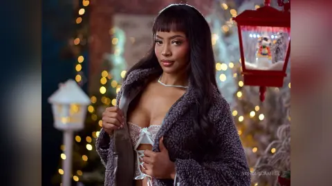 NohomiCambel wears a thick grey fuzzy coat over a white lace-up bra, posing against a soft-focus background of warm holiday lights.