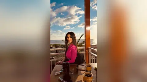 JuliDeming sits at an outdoor seaside cafe table wearing a pink long-sleeve top under a bright blue sky.