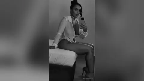 Glamorous woman in a sheer white blouse and briefs sitting on a bed in a moody black and white portrait.