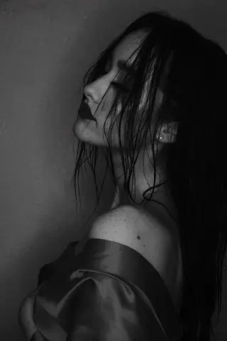 Moody black and white portrait of a woman with wet hair in a satin dress against a dark wall.