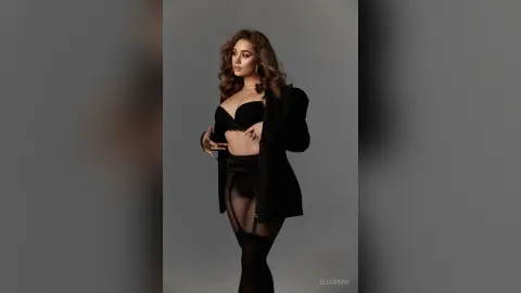 Elegant woman in black lingerie and blazer posing confidently against a grey studio background.