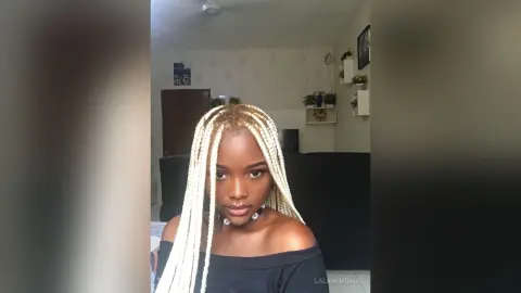 Young woman with long blonde box braids wearing a black off-the-shoulder top in a bright indoor setting.